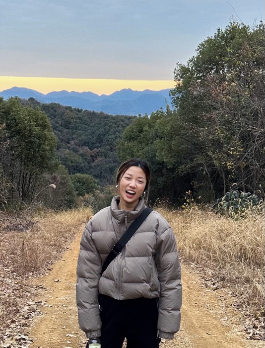 Maxine Zhou outdoors on a hillside trail at dusk, laughing, with wooded mountains behind her